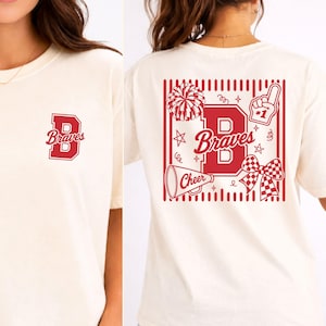 Op de afbeelding: Een off-white t-shirt met een rode afbeelding op de linkerborst met een grote letter B en het woord Braves. De achterkant toont een grote rode afbeelding met een letter B, het woord Braves, een pompon, een megafoon, een strik en een schuimvinger.