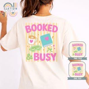May include: Cream-colored t-shirt with a graphic that reads "BOOKED & BUSY" in pink and green. The design includes a coffee cup, book, eye mask, and other elements on a striped background.