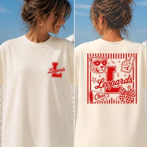 Op de afbeelding: Een crèmekleurig T-shirt met een kleine rode letter L en het woord Leopards op de linkerborst. De achterkant toont een grote rode afbeelding met een luipaard met een zonnebril, een megafoon, een schuimvinger en een geruite strik.
