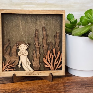 May include: A wooden shadow box featuring a carved axolotl, plants, and the inscription "Starburst" at the base. The box is displayed on a wooden surface.