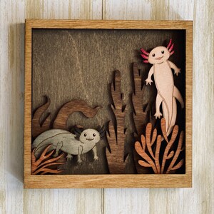 May include: A wooden wall art piece featuring a brown background with a cutout of two axolotls, one pink and one grey, surrounded by brown and orange cutout plants.