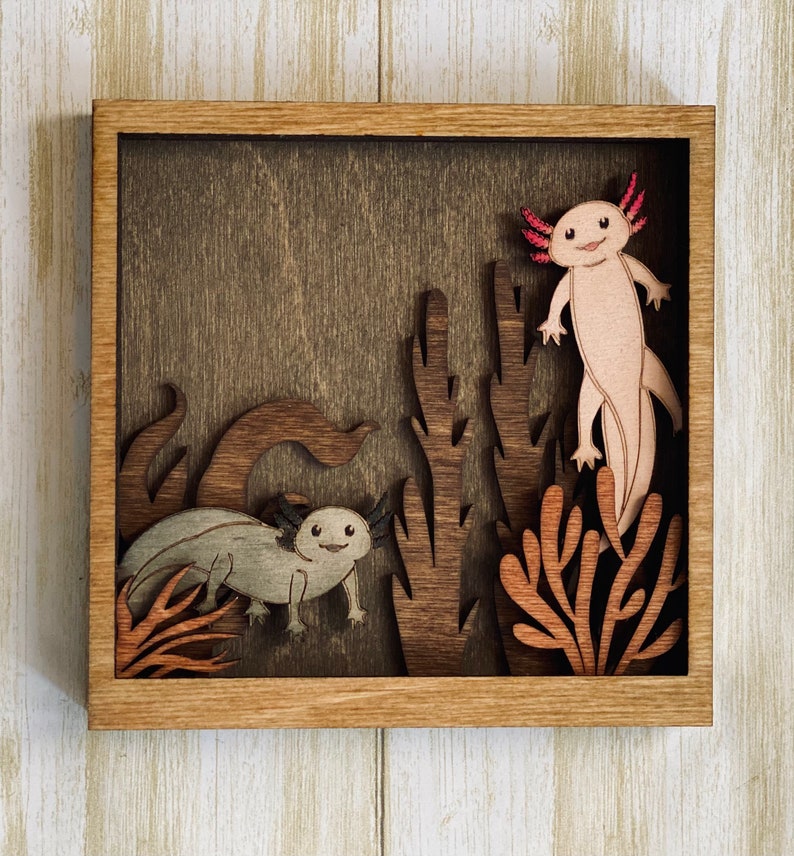 May include: A wooden wall art piece featuring a brown background with a cutout of two axolotls, one pink and one grey, surrounded by brown and orange cutout plants.