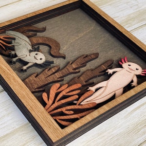 May include: A wooden framed shadow box with a 3D layered design of two axolotls in a coral reef setting. The axolotls are carved from wood and painted in shades of pink and grey. The coral reef is made of brown and orange wood.