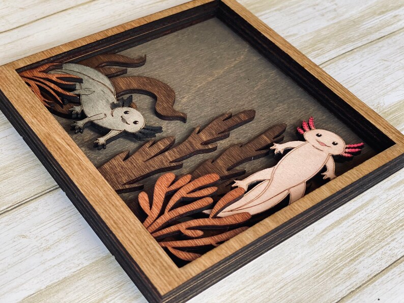 May include: A wooden framed shadow box with a 3D layered design of two axolotls in a coral reef setting. The axolotls are carved from wood and painted in shades of pink and grey. The coral reef is made of brown and orange wood.