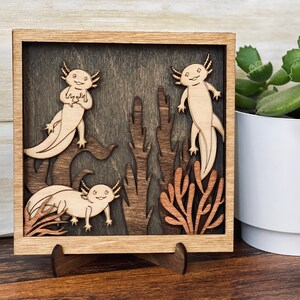 May include: A wooden wall art piece featuring three axolotls in a dark brown frame. The axolotls are carved in a light brown colour and are surrounded by brown and orange coral-like plants.