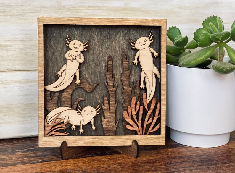 May include: A wooden wall art piece featuring three axolotls in a dark brown frame. The axolotls are carved in a light brown colour and are surrounded by brown and orange coral-like plants.