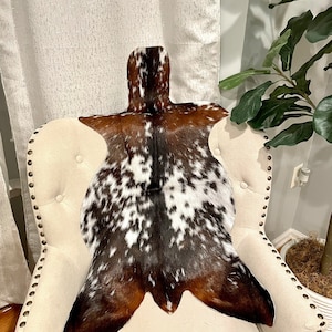 Puede incluir: Una alfombra de piel de vaca marrón y blanca con un patrón de manchas. La alfombra está colocada sobre un sillón blanco con tapicería capitoné.