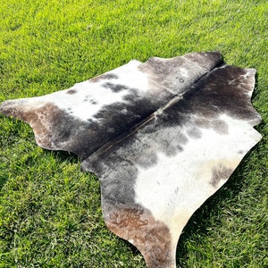 May include: A white and brown cowhide rug with a natural, irregular shape. The rug is laid out on a green grassy surface.