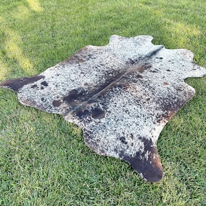 Pode incluir: Um tapete de pele de vaca preto e branco com um padr&atilde;o natural manchado. O tapete est&aacute; colocado sobre uma superf&iacute;cie de grama verde.
