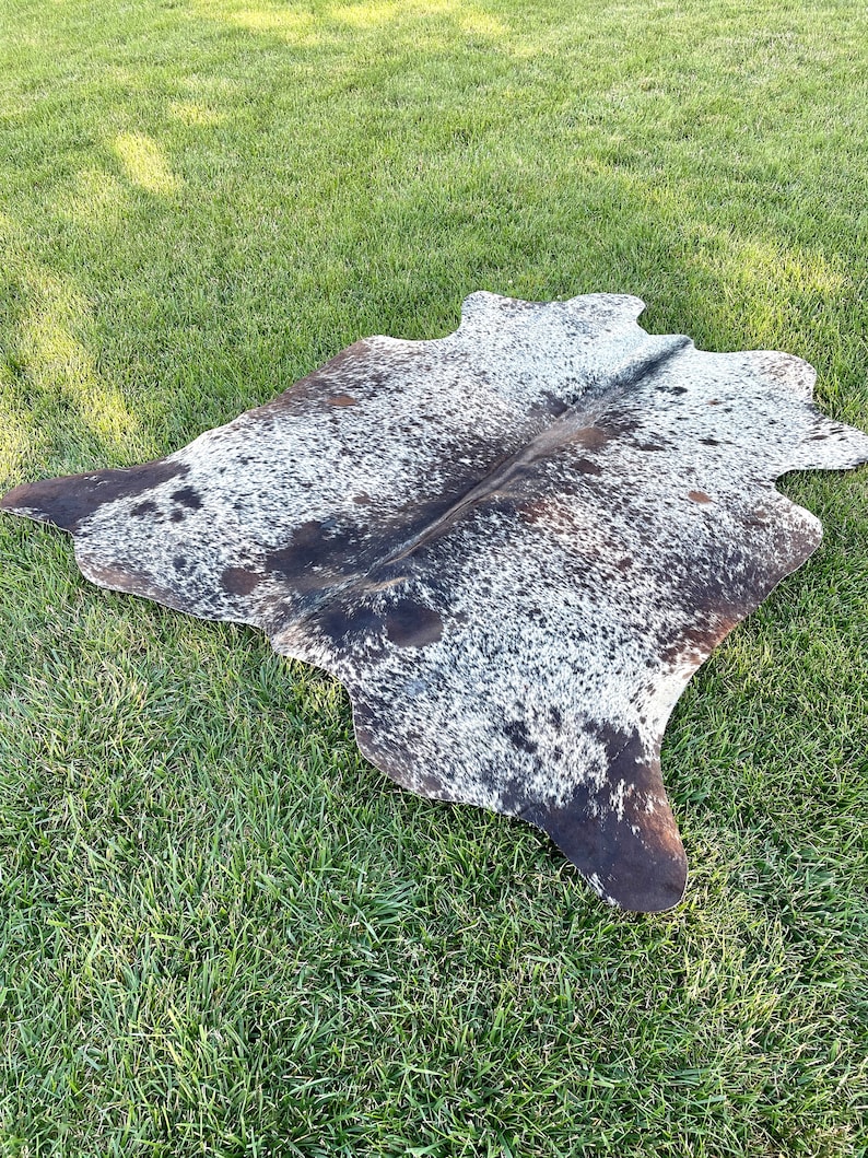Pode incluir: Um tapete de pele de vaca preto e branco com um padr&atilde;o natural manchado. O tapete est&aacute; colocado sobre uma superf&iacute;cie de grama verde.