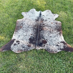 Pode incluir: Um tapete de pele de vaca branco e marrom com um padr&atilde;o de manchas. O tapete est&aacute; colocado sobre uma superf&iacute;cie de grama verde.