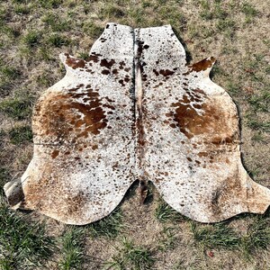 Pode incluir: Um tapete de pele de vaca marrom e branco com um padr&atilde;o de manchas. O tapete est&aacute; colocado sobre uma superf&iacute;cie gramada.