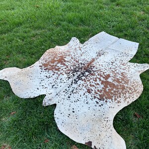 Pode incluir: Um tapete de pele de vaca branco com manchas marrons e pretas. O tapete est&aacute; colocado sobre uma superf&iacute;cie de grama verde.