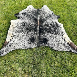 K&ouml;nnte beinhalten: Ein schwarz-wei&szlig;er Kuhfellteppich mit einem gefleckten Muster. Der Teppich liegt auf einer gr&uuml;nen Grasfl&auml;che.