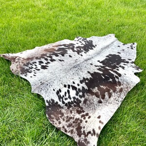 Pode incluir: Um tapete de pele de vaca preto e branco com manchas marrons. O tapete est&aacute; colocado sobre uma superf&iacute;cie de grama verde.