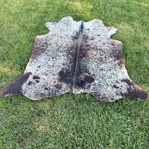 Pode incluir: Um tapete de pele de vaca marrom e branco com um padr&atilde;o de manchas. O tapete est&aacute; colocado sobre uma superf&iacute;cie de grama verde.