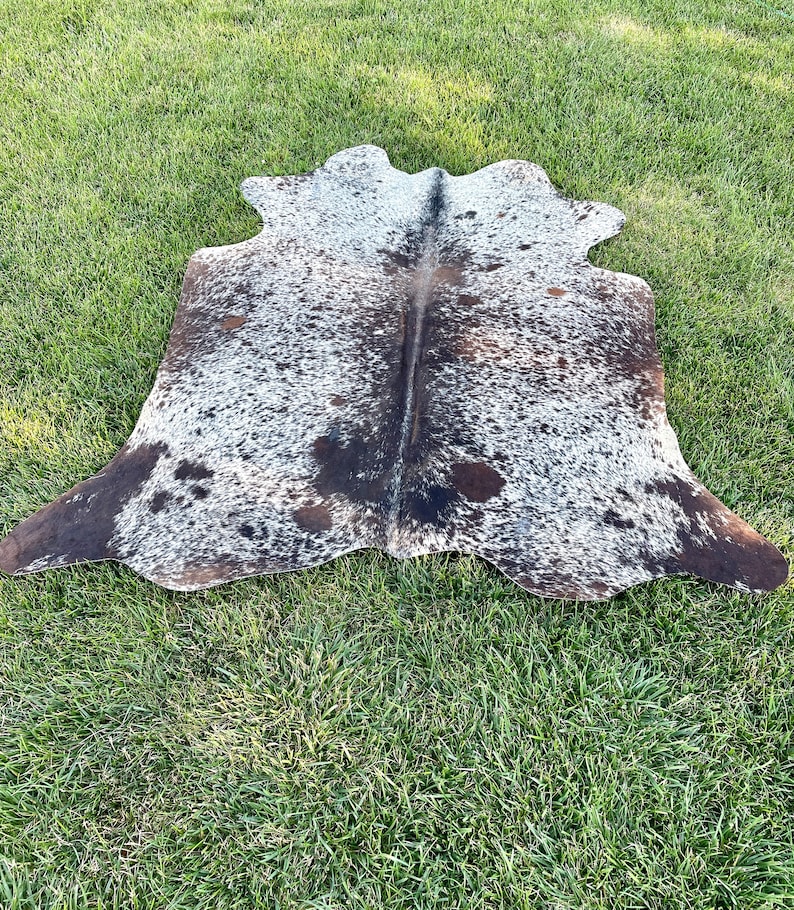 Pode incluir: Um tapete de pele de vaca marrom e branco com um padr&atilde;o de manchas. O tapete est&aacute; colocado sobre uma superf&iacute;cie de grama verde.