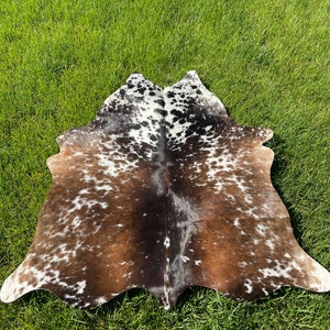 May include: A brown and white cowhide rug with a spotted pattern. The rug is laid out on a green grassy surface.