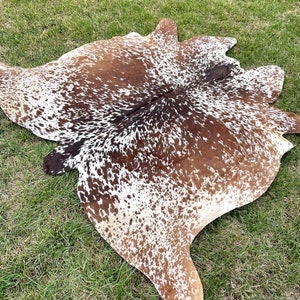 May include: A brown and white cowhide rug with a spotted pattern. The rug is laid out on a green grassy surface.
