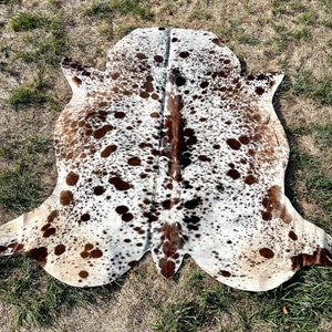 Pode incluir: Um tapete de pele de vaca marrom e branco com um padr&atilde;o de manchas. O tapete est&aacute; colocado sobre uma superf&iacute;cie coberta de grama.