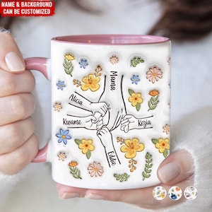 May include: White ceramic mug with a pink rim, featuring a design of five hands holding each other with the names "Mama", "Alicia", "Kwame", "Kezia", and "Eddie" written on the fingers. The mug is surrounded by a floral pattern with yellow, orange, and blue flowers.