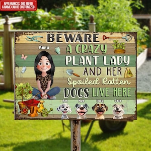 Op de afbeelding: Een groen en bruin metalen bord met de tekst "Pas op voor een gekke plantenlady en haar verwende, rotte honden die hier wonen". Het bord toont een cartoonvrouw met bruin haar en een groen shirt, en vier cartoonhonden. Het bord is perfect voor een tuin of erf.