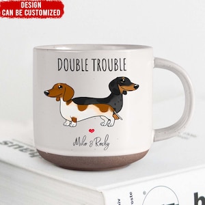 May include: White ceramic mug with a brown base. The mug features a cartoon illustration of two dachshund dogs with the text "Double Trouble" above them and "Milo & Rocky" below them.