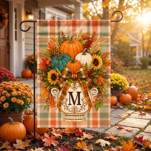 May include: A rectangular garden flag featuring a plaid background in muted orange, green, and cream. The design displays a decorative urn with the letter M, overflowing with pumpkins, sunflowers, wheat stalks, and autumn leaves, tied with a plaid bow.