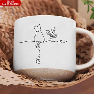 May include: White ceramic mug with a brown base, featuring a continuous line drawing of a cat, holly, and the name "Olivia". The mug has a curved handle and is set in a brown paper-filled box.