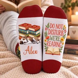 May include: A pair of white socks with red cuffs featuring a sleeping beagle, a stack of colourful books, and the name Alex on one sock, and the text DO NOT DISTURB WE'RE READING with floral accents and an open book graphic on the other sock.