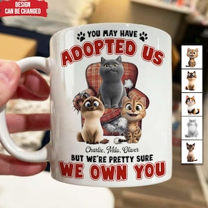 May include: White ceramic mug with a handle featuring a cartoon image of three cats sitting on a plaid armchair. The text reads "YOU MAY HAVE ADOPTED US, BUT WE'RE PRETTY SURE WE OWN YOU." The names Charlie, Milo, and Oliver are also printed on the mug.