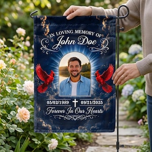 Può includere: Una bandiera da giardino rettangolare con sfondo cielo blu scuro, nuvole e due cardinali rossi. Il testo recita IN LOVING MEMORY OF John Doe, 05/02/1989 - 09/21/2025, e Forever In Our Hearts. Un arco centrale mostra la foto di un uomo.