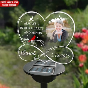 May include: A heart-shaped memorial garden stake with a photo, the text "ALWAYS IN OUR HEARTS AND MINDS...", a red cardinal, and the name "Sarah" with a date. The stake is solar-powered.