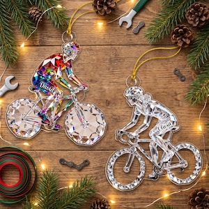 Può includere: Due ornamenti in acrilico a forma di ciclisti su biciclette. Un ornamento presenta un motivo geometrico multicolore con il nome David stampato sul telaio. L'altro ornamento è trasparente con un design geometrico e il nome Olivia sul telaio.
