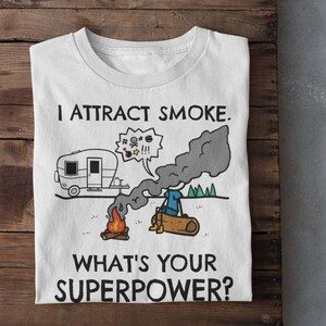 May include: White t-shirt with a cartoon illustration of a person sitting by a campfire with a camper van in the background. The text on the shirt reads "I attract smoke. What's your superpower?"