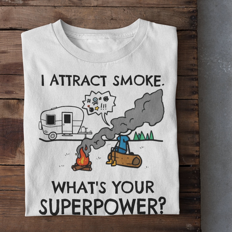 May include: White t-shirt with a cartoon illustration of a person sitting by a campfire with a camper van in the background. The text on the shirt reads "I attract smoke. What's your superpower?"