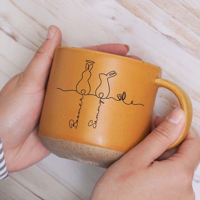 May include: Mustard yellow ceramic mug featuring a minimalist black line drawing of two rabbits and the names "Boomer" and "Sammy". The mug has a light brown base and a curved handle.
