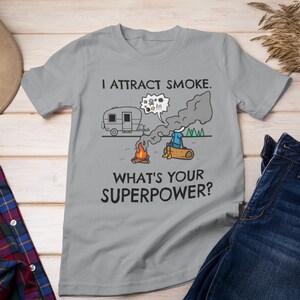 May include: A gray t-shirt with the text "I attract smoke. What's your superpower?" and a cartoon illustration of a camper, a campfire, and a person with smoke coming from their head.
