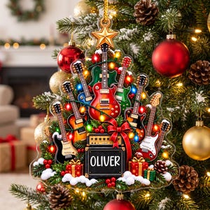 Puede incluir: Un adorno con forma de árbol de Navidad que presenta varias guitarras eléctricas en rojo, azul, verde y negro, dispuestas alrededor de un amplificador central con el nombre OLIVER impreso. El diseño incluye una estrella dorada, lazos rojos y regalos.