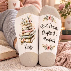 May include: A pair of beige and white socks. The left sock features a stack of illustrated books topped with flowers and the name Emily. The right sock displays the text Quiet Please, Chapter In Progress with floral accents.