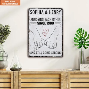 以下が含まれることがあります： 「SOPHIA & HENRY」と「ANNOYING EACH OTHER SINCE 1980 AND STILL GOING STRONG」の文字が入った長方形の金属製の看板。赤いハートで互いを抱き合う2つの手のグラフィックが特徴です。看板は、ヴィンテージ感のある外観です。