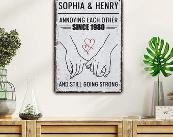 Könnte beinhalten: Rechteckiges Metallschild mit dem Text "SOPHIA & HENRY" und "ANNOYING EACH OTHER SINCE 1980 AND STILL GOING STRONG". Das Schild zeigt eine Grafik von zwei Händen, die sich mit einem roten Herzen halten. Das Schild hat einen Vintage-Look.