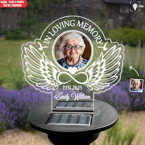 May include: A memorial solar light with a photo of a woman, the text "IN LOVING MEMORY", the years 1951-2025, and the name "Emily William". The design includes angel wings and an infinity symbol. The light is on a black base.