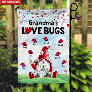 May include: A garden flag with a gnome wearing a red and white hat and holding a flower. The flag says "Grandma's Love Bugs" and has the names of family members around the gnome.