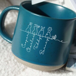May include: Teal ceramic mug featuring a white line drawing of bunnies and names: Cooper, Charles, Teddy, and Barney. The mug has a speckled finish and a light beige base.