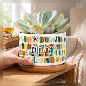 Può includere: Un vaso cilindrico in ceramica bianca con una colorata illustrazione di una libreria piena di libri e motivi floreali. Il nome Olivia è stampato in grandi lettere multicolori sulla parte anteriore. Il vaso poggia su una base rotonda in legno e contiene una grande pianta succulenta verde.