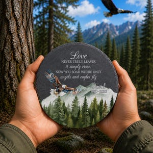 May include: A circular slate memorial plaque featuring a printed illustration of a bald eagle flying over a mountain range and pine forest. The text reads: Love NEVER TRULY LEAVES it simply rises. NOW YOU SOAR WHERE ONLY angels and eagles fly David 1960-2026.