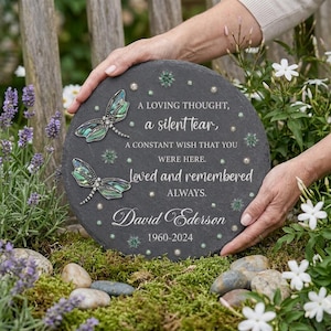 Op de afbeelding: Een ronde gedenkplaat van leisteen met twee iriserende libelle-illustraties en de tekst: A LOVING THOUGHT, a silent tear, A CONSTANT WISH THAT YOU WERE HERE. Loved and remembered ALWAYS. David Ederson 1960-2024, versierd met kleine zilveren stippen.