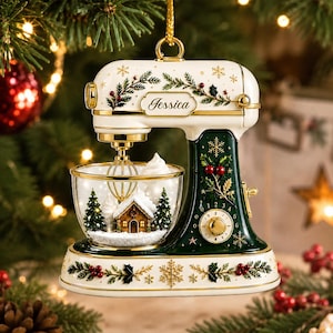 Peut inclure: Une décoration de Noël en forme de robot pâtissier crème et vert foncé avec des accents dorés, des motifs de houx et de baies, et le prénom Jessica inscrit sur le devant. Le bol contient une maison enneigée et des sapins miniatures.