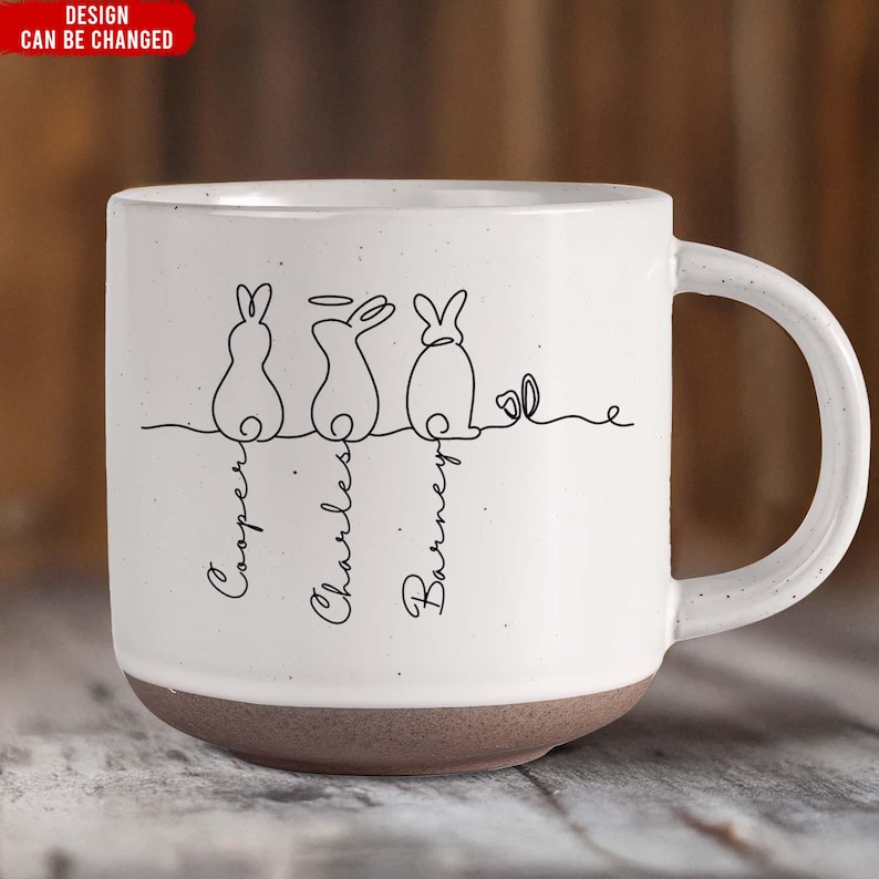 May include: White ceramic mug with a brown base. The mug features a line drawing of three bunnies with names below them. The text "DESIGN CAN BE CHANGED" is in a red rectangle in the top left corner.
